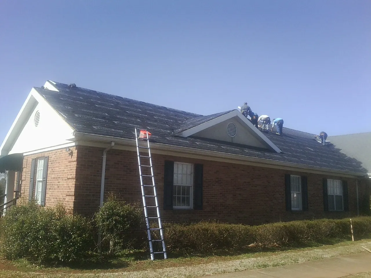 Crew tearing shingles off a brick ranch roof with a ladder set — Emergency Roof Repair work by our crew in Hanahan