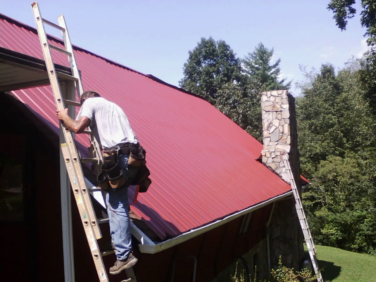 Roofer on a ladder beside a red metal roof and stone chimney — Soffit & Fascia Repair by Roofing Brothers in Hanahan